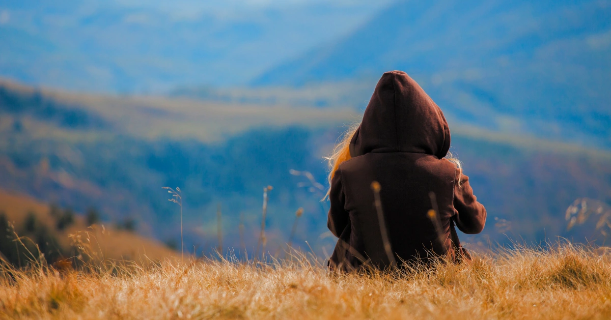 a girl sitting on a hill look out at the mountains
